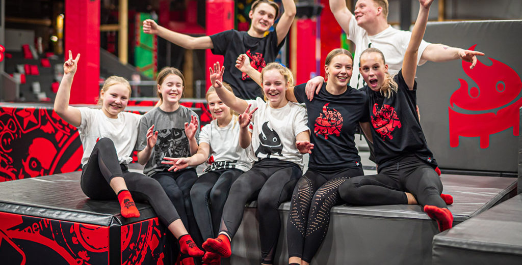 Yngre tonåringar och barn sitter uppradade för foto på JumpYard. De vill träna i Göteborg! De har tröjor med JumpYard-logga på sig, Alla har armarna upp i luften och ser glada ut.
